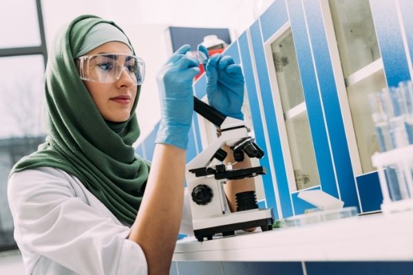 A woman wearing a headscarf and protective goggles works at the microscope