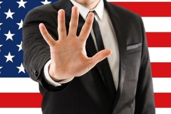 A man stands in front of a US-American flag and holds his hand defensively into the camera